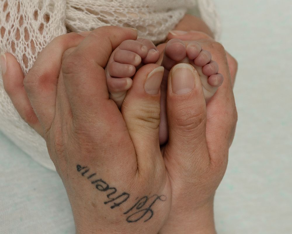 Adult hands cradling tiny newborn baby feet, with a cursive tattoo visible on the wrist.