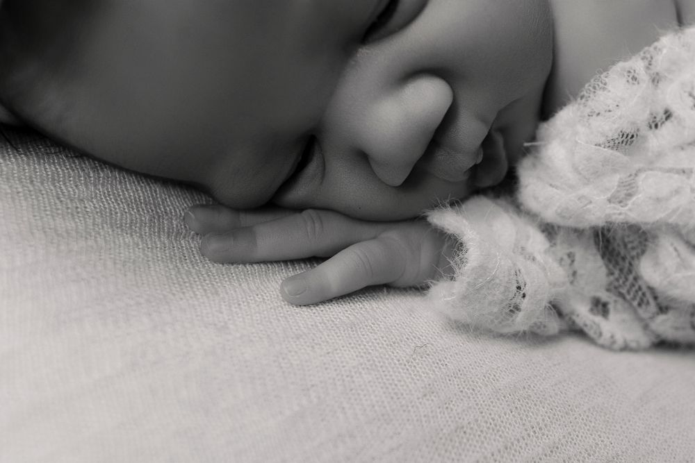 Black and white close-up of a sleeping newborn baby with tiny fingers resting near a soft lace toy.
