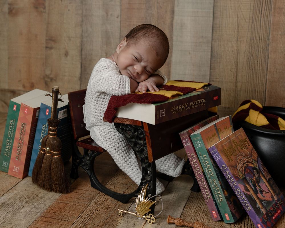 Baby dressed in white sits at a tiny desk surrounded by Harry Potter books, broom, and Gryffindor accessories.