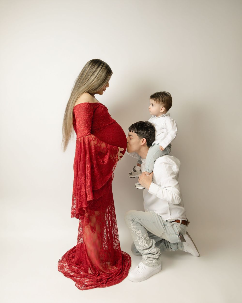 Pregnant woman in red lace gown with partner kneeling kissing belly and toddler sitting on shoulders.
