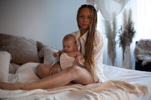 en mamma sitter på sängen med sitt barn i knät. mamman kollar in i kameran. bakgrunden är ljus