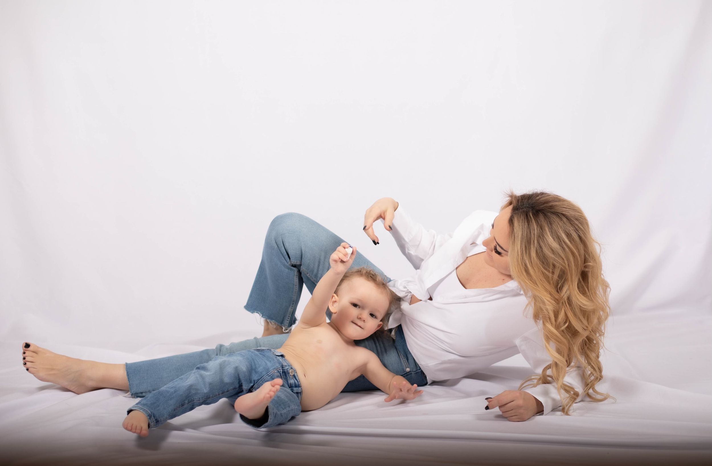 mamma och ett barn ligger på golvet i bakgrunden syns tyg. båda bär jeans, mamman kollar på barnet och barnet kollar mot kameran.
