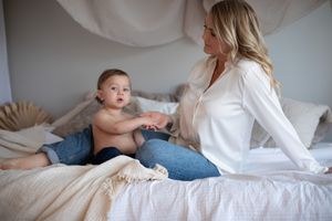 kvinna sitter i en säng iklädd en vit skjorta och ljusa jeans. framför sig har hon sitt barn som hon håller i handen.