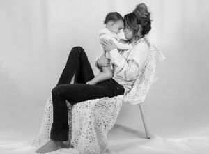 kvinna sitter på en stol iklädd tyg. hon håller upp sitt barn emot sig och de har pannorna emot varandra. kvinnan ler.