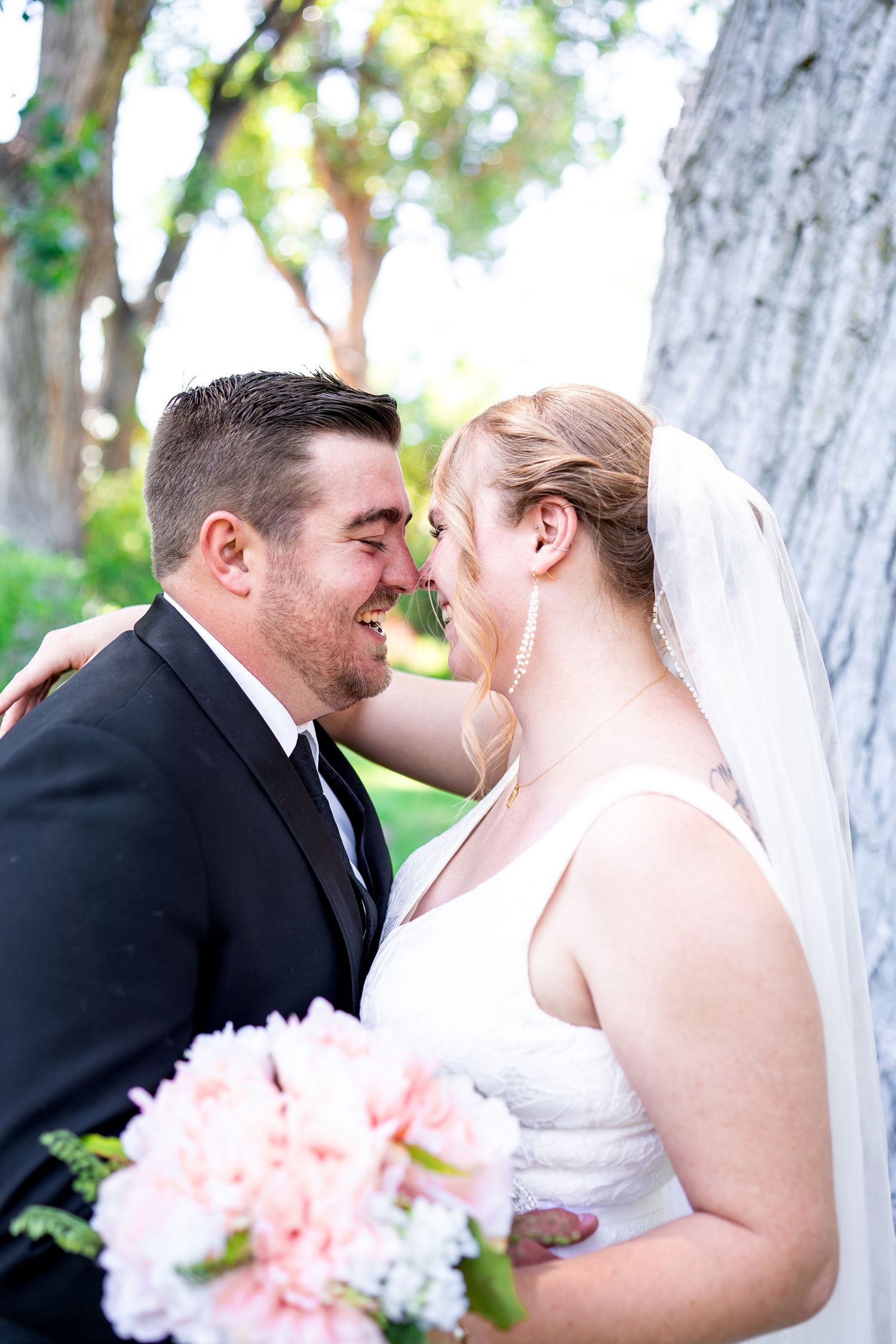 A tender moment between newlyweds with pink bouquet against a tree trunk in natural lighting.