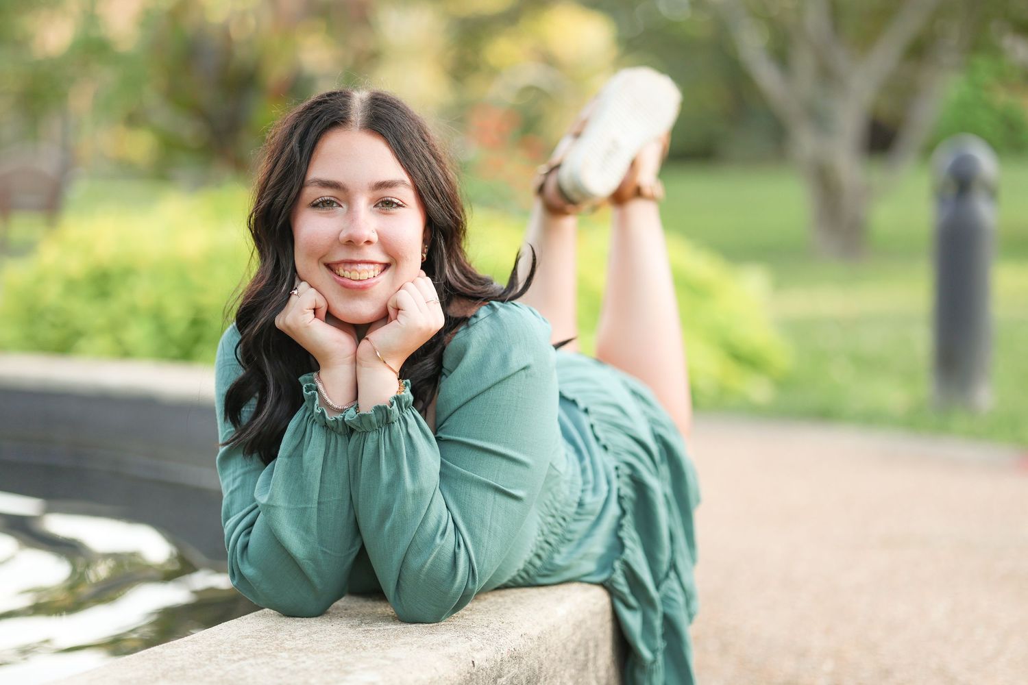 A person in teal dress poses while lying on stone wall outdoors with autumn scenery.