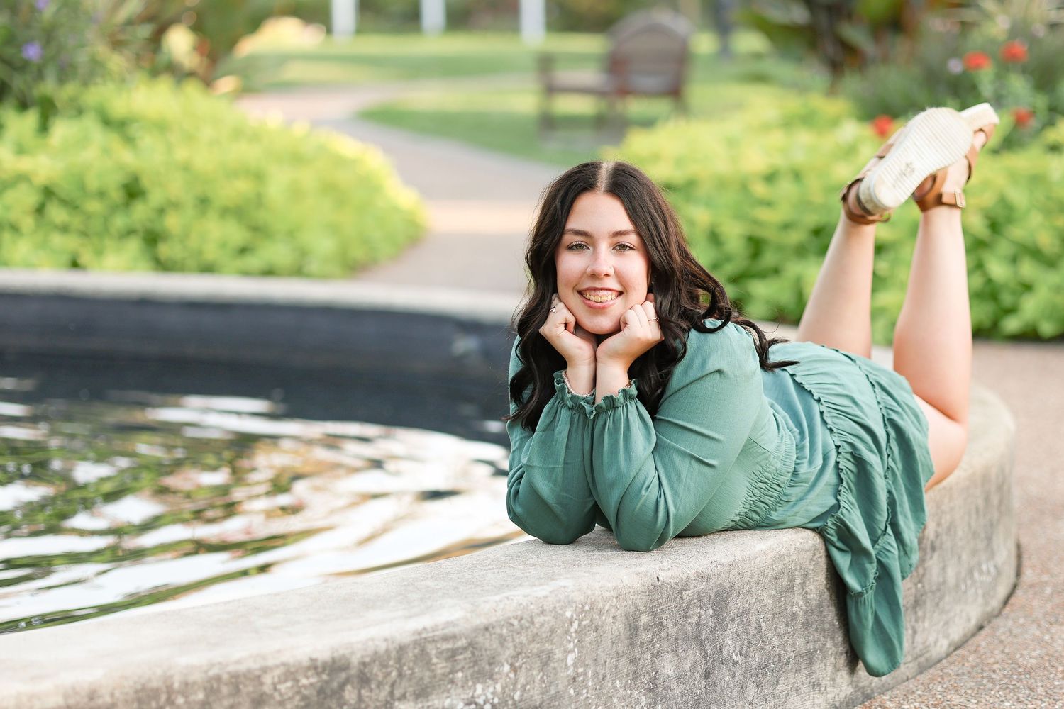 A person in teal dress poses playfully on stone wall near water feature in garden setting.