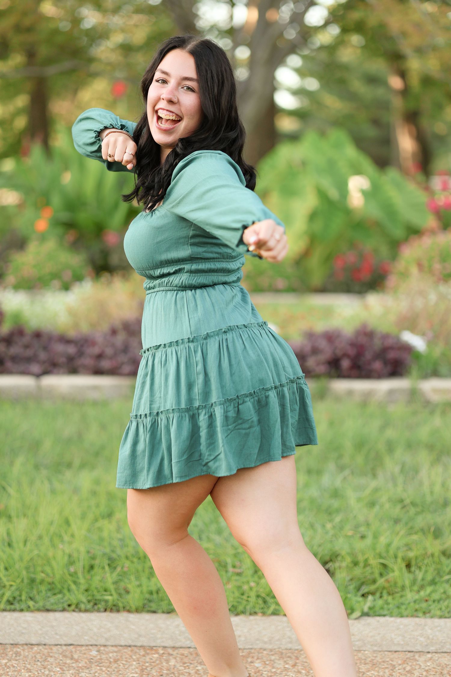 A person in teal dress dances energetically outdoors in a garden setting.