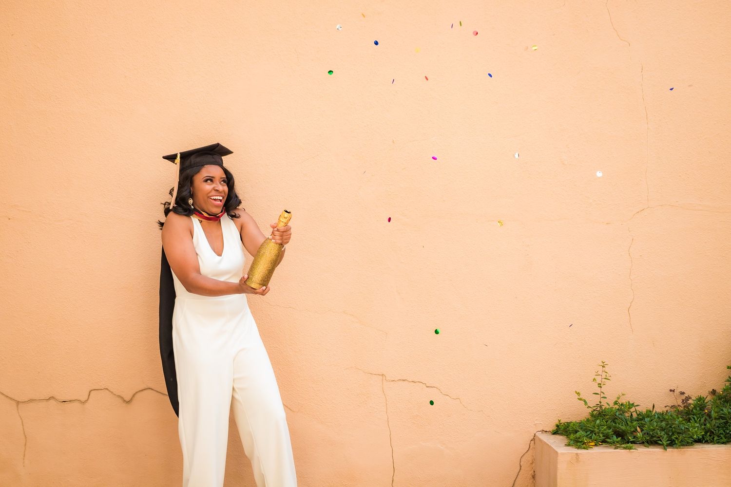 Graduate in white jumpsuit celebrating with champagne bottle against peach colored wall.