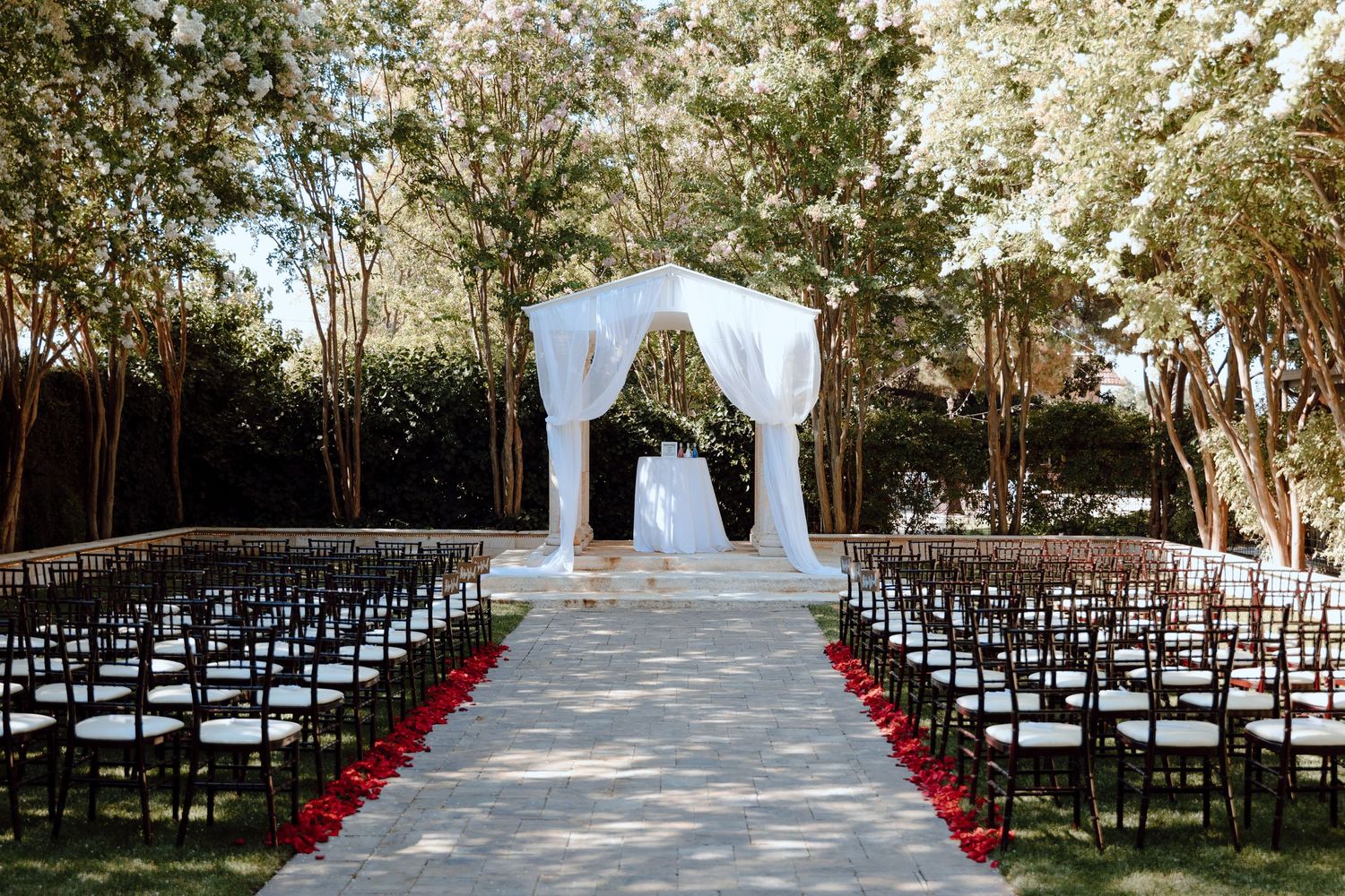Outdoor wedding ceremony setup with white arch and rows of dark chairs along rose petal aisle.