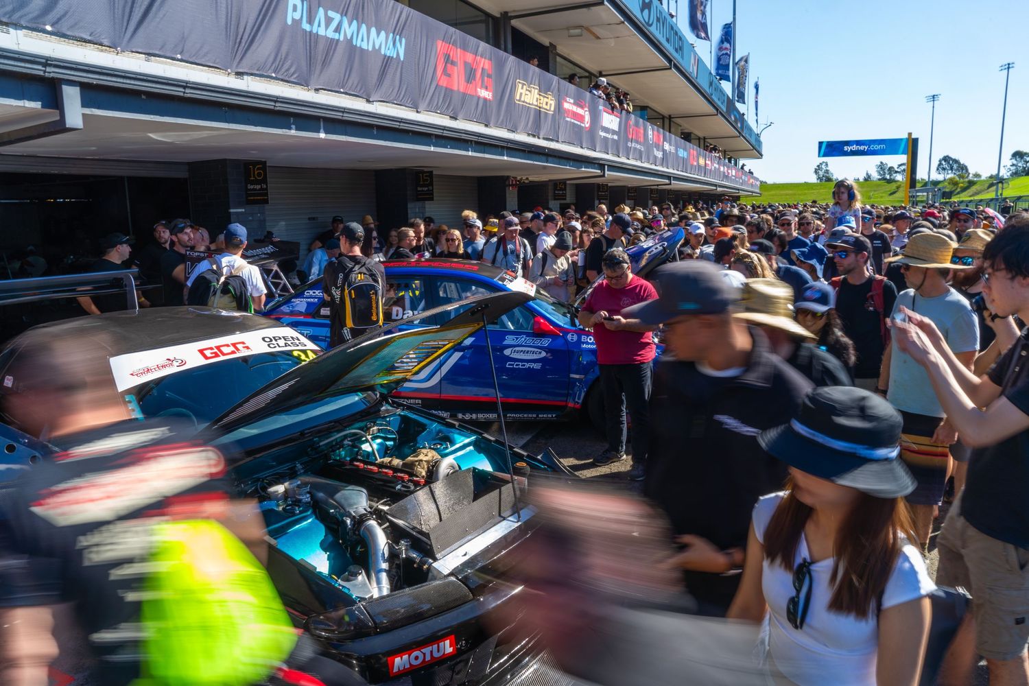 Crowded racing pit lane with fans gathering around a Formula-style race car.