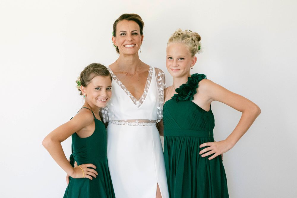 A bride in a white lace dress poses with two attendants wearing emerald green gowns against a light backdrop.