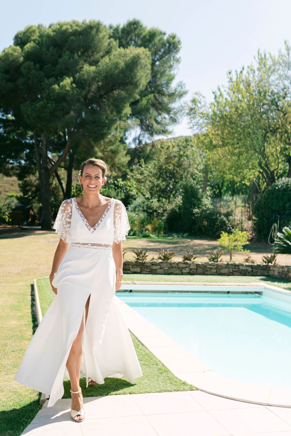A bride in a lace wedding dress poses by a swimming pool in a sunny garden setting.