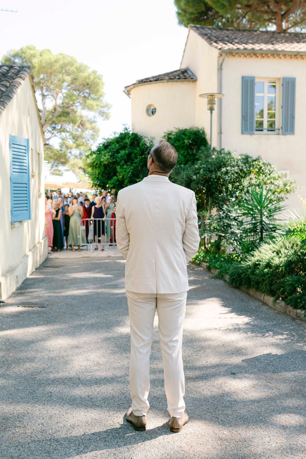 A person in a cream colored suit stands facing away from the camera with guests visible in the background.