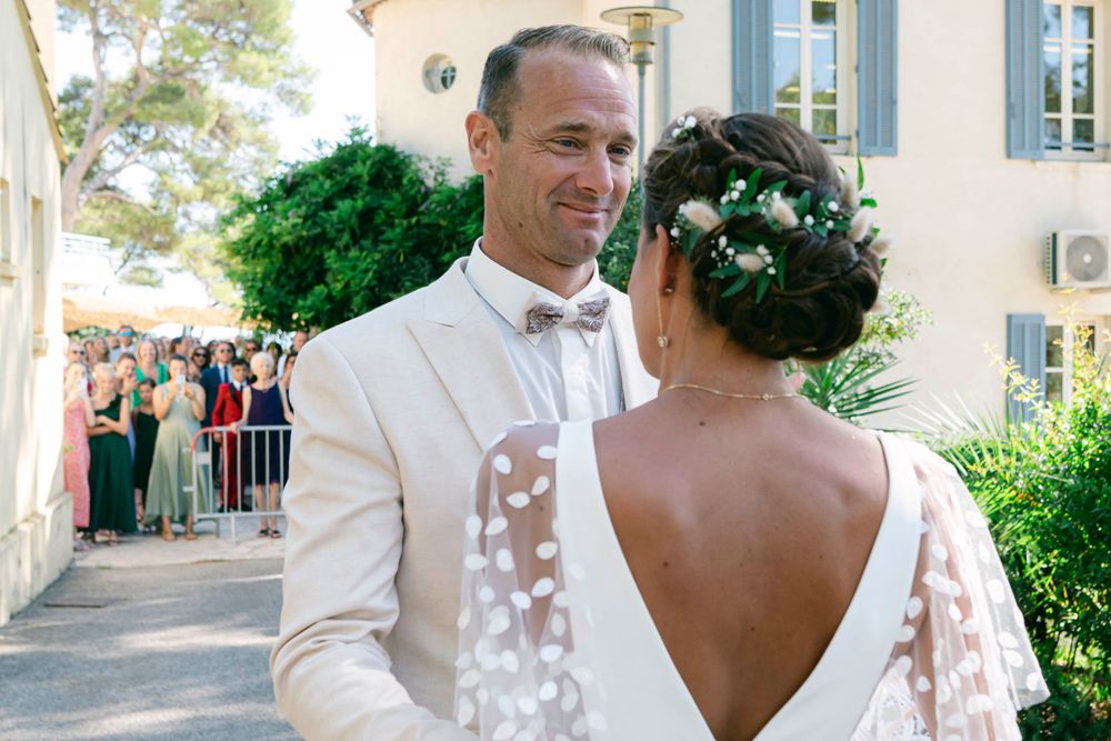A wedding couple shares an intimate moment outdoors with guests visible in the background.