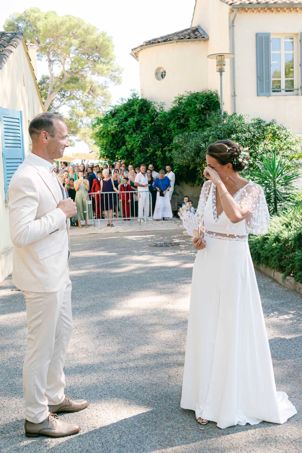 A wedding couple stands facing each other on a path with guests watching in the background.