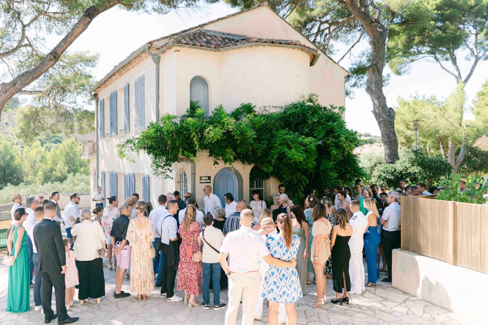 Wedding guests gather outside a Mediterranean-style venue with blue shutters and stone walls.