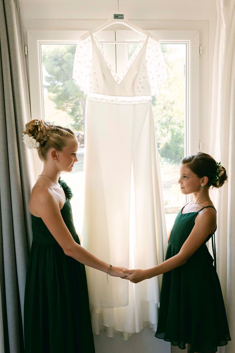 Two bridesmaids in dark green dresses standing beside a hanging white wedding gown near a window.