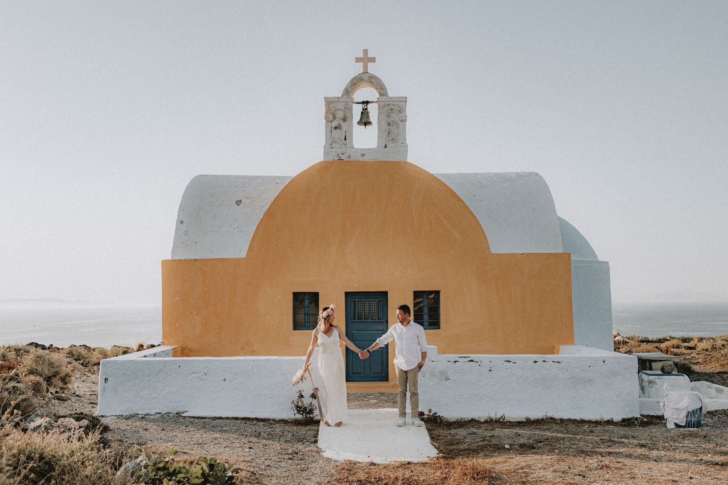 Bride and groom holding hands in front of orange and white chapel in Santorini