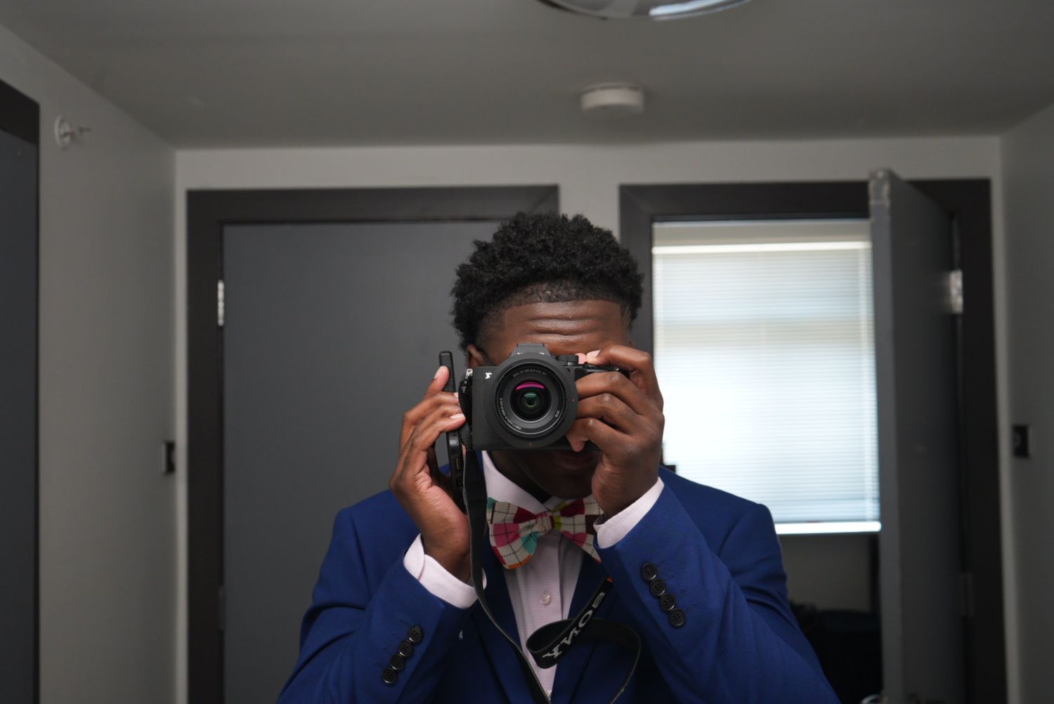 A man in a blue suit and floral bow tie takes a mirror selfie, holding a black camera in front of his face.