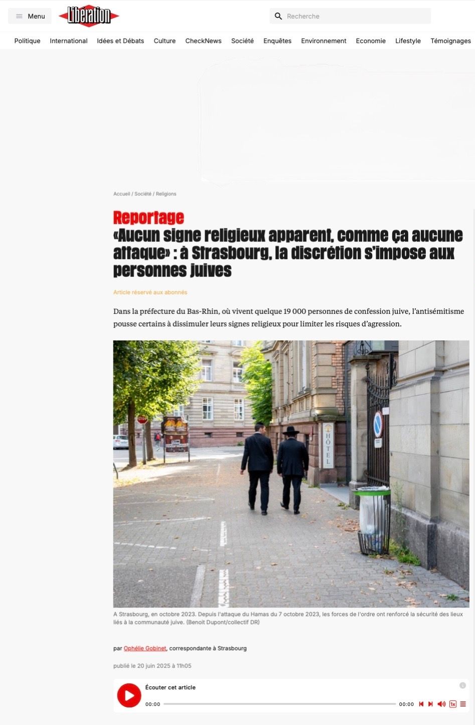 Two figures walking down a quiet European city street with buildings and trees on a sunny day.