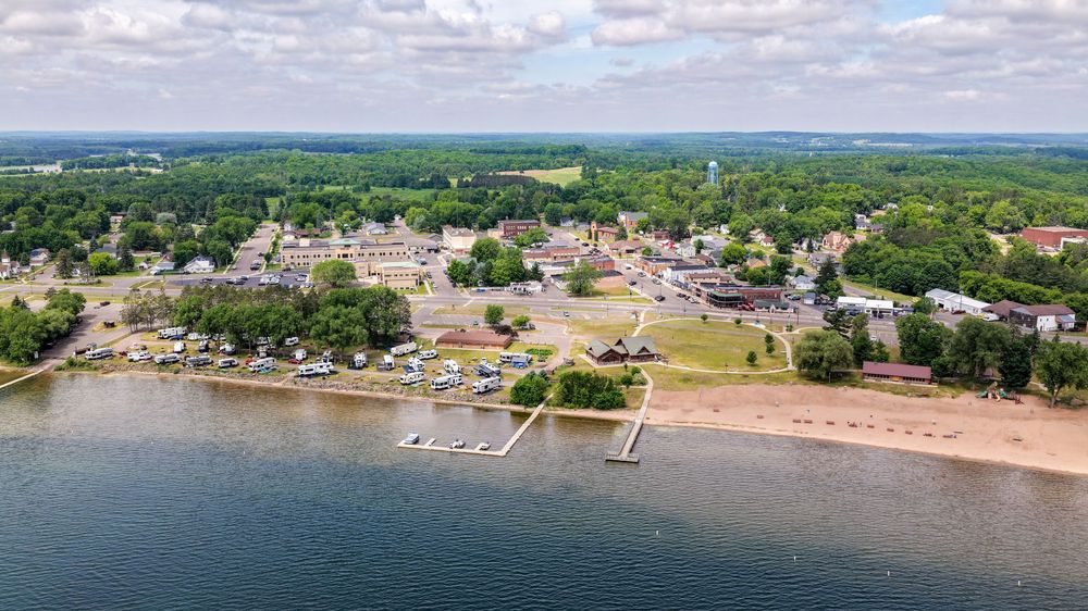 Aerial view of a small lakeside town with sandy beach, marina docks, lush trees, and buildings under a partly cloudy sky.