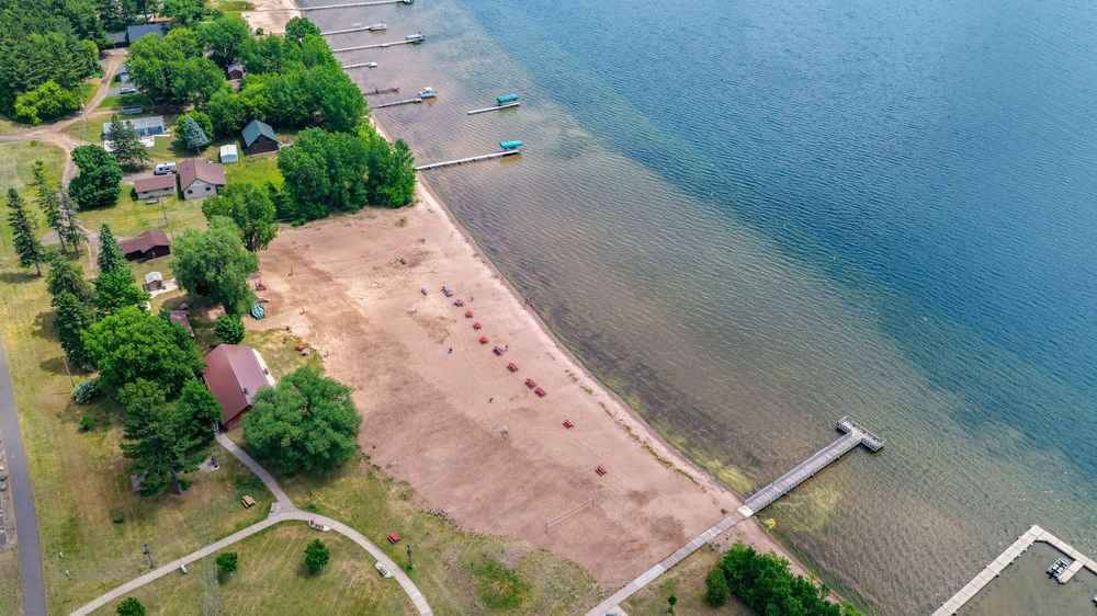 Aerial view of a sandy lakeside beach with a dock, green trees, and calm blue water on a sunny day.