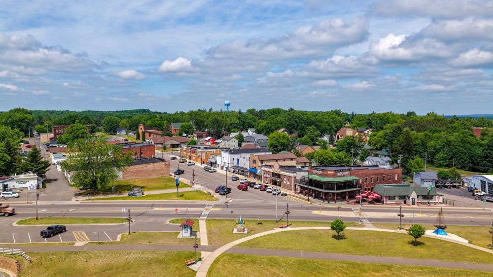 Aerial view of a small town with brick buildings, tree-lined streets, and an intersection under a partly cloudy sky.