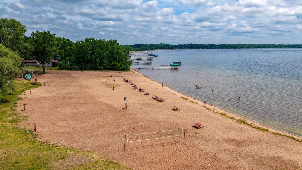 Aerial view of a sandy beach on a calm lake with volleyball nets, boats, and lush green trees under a cloudy sky.