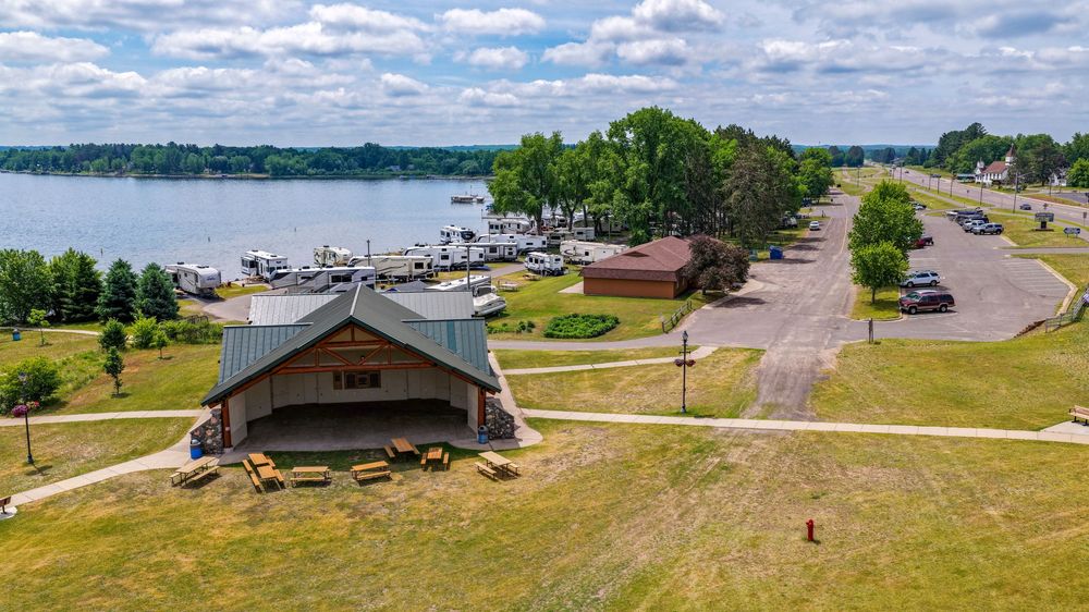 Aerial view of a lakeside marina with docked boats, a pavilion, green lawns, and a tree-lined road on a sunny day.