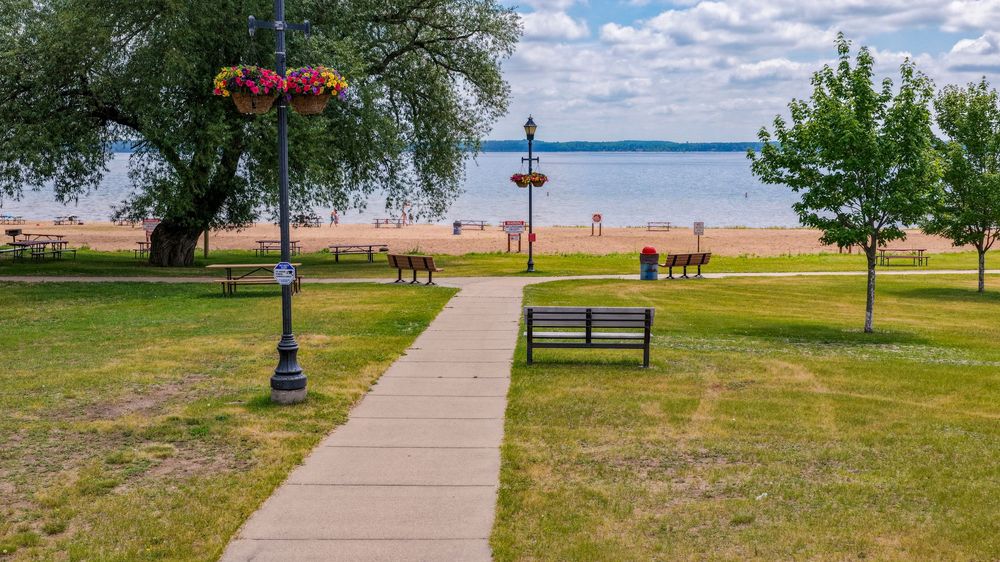 A peaceful park pathway lined with benches and flower-hanging lamp posts leading to a sandy lake beach.