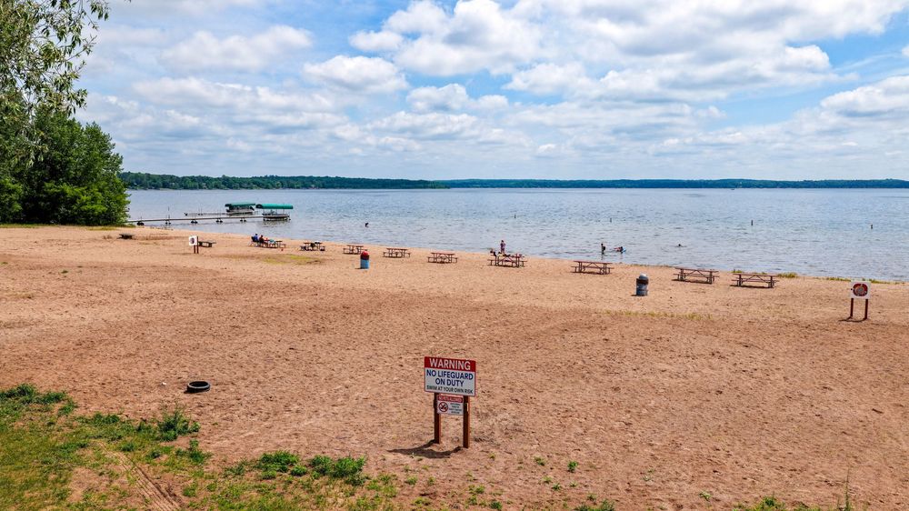 Sandy lakeside beach with picnic benches, a warning sign, calm water, and partly cloudy skies on a quiet day.