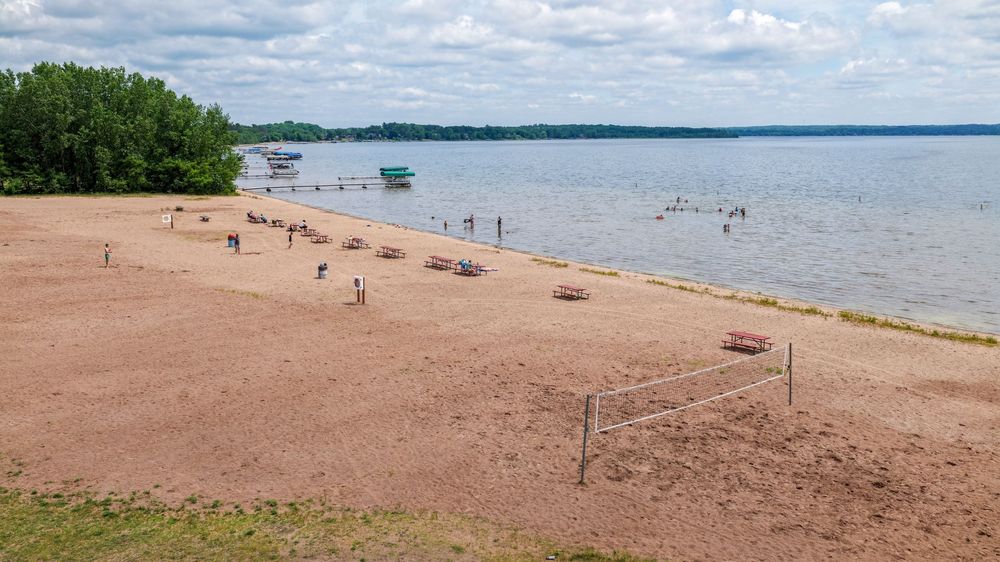 Sandy beach on a lake with volleyball net, scattered visitors, docks, and partly cloudy skies overhead.