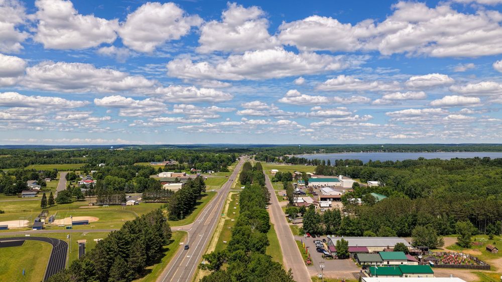 Aerial view of small town highway with lake and lush green trees under cloudy blue sky.