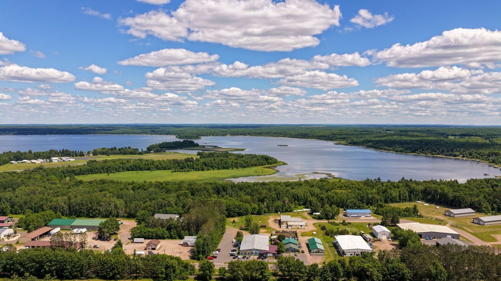 Aerial view of a forested lakeside community with buildings nestled among trees beside a wide river under blue skies.