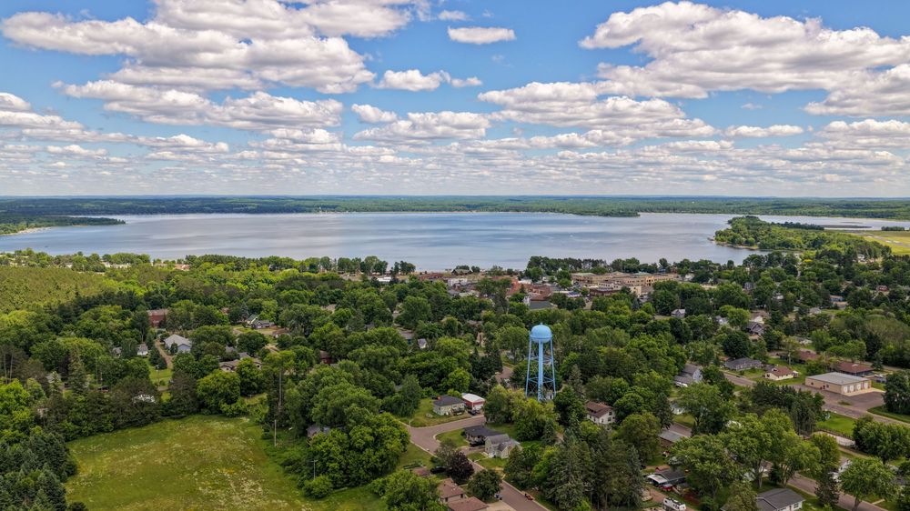 Aerial view of a quiet residential town surrounded by lush trees with a blue water tower and large lake beyond.