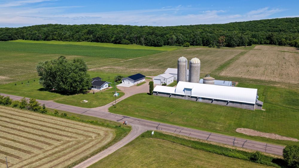Aerial view of a rural farm with silos, green fields, and a large white barn surrounded by lush trees and cropland.