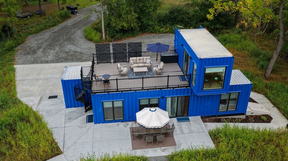 Aerial view of a modern blue shipping container home with rooftop solar panels, outdoor patio furniture, and a paved terrace.