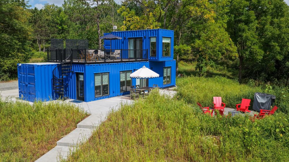 Blue shipping container home with rooftop deck, patio umbrella, and red chairs surrounded by lush green trees and meadow.