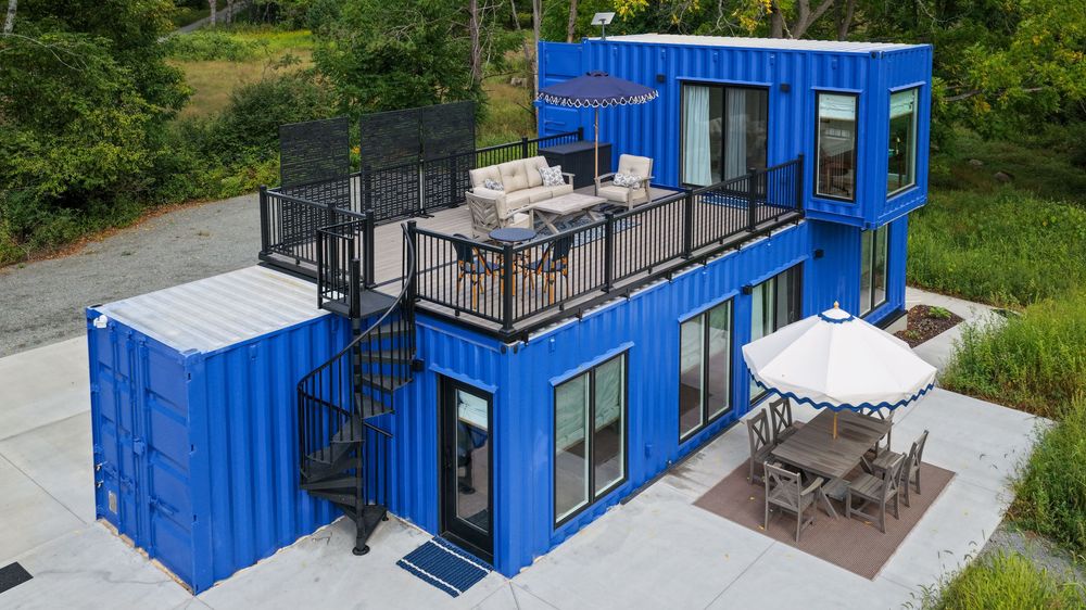 Blue shipping container home with rooftop deck, spiral staircase, outdoor dining area, and modern design surrounded by lush greenery.