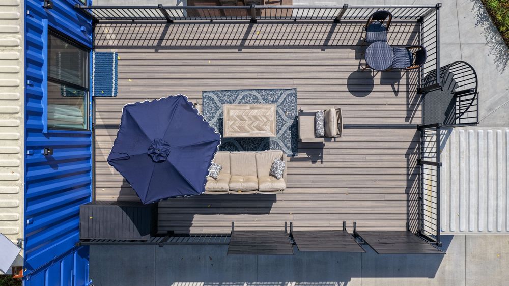 Aerial view of a rooftop deck with outdoor seating, blue patio umbrella, and bistro table set on gray composite decking.