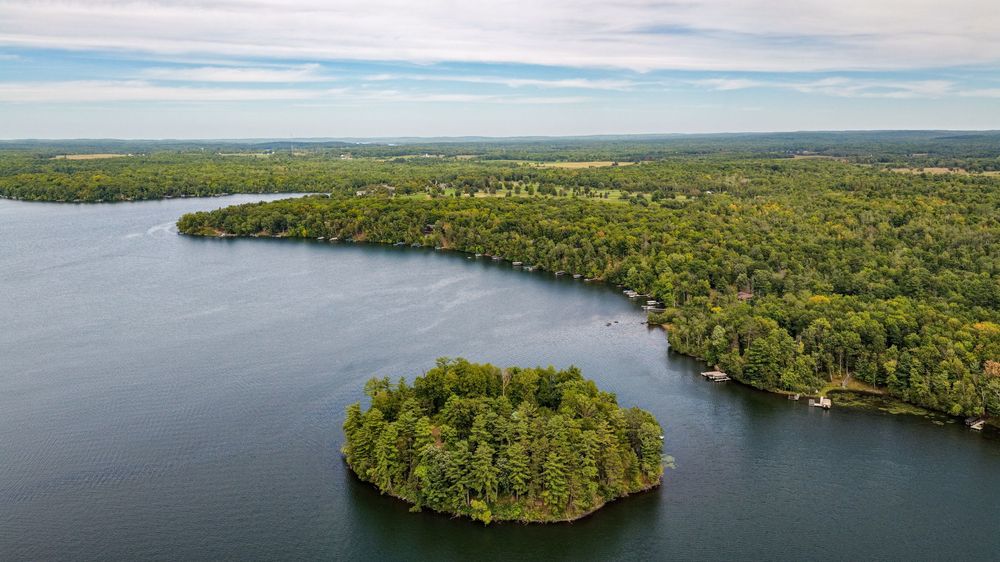 Aerial view of a lush green island and forested peninsula surrounded by calm blue lake waters under a cloudy sky.