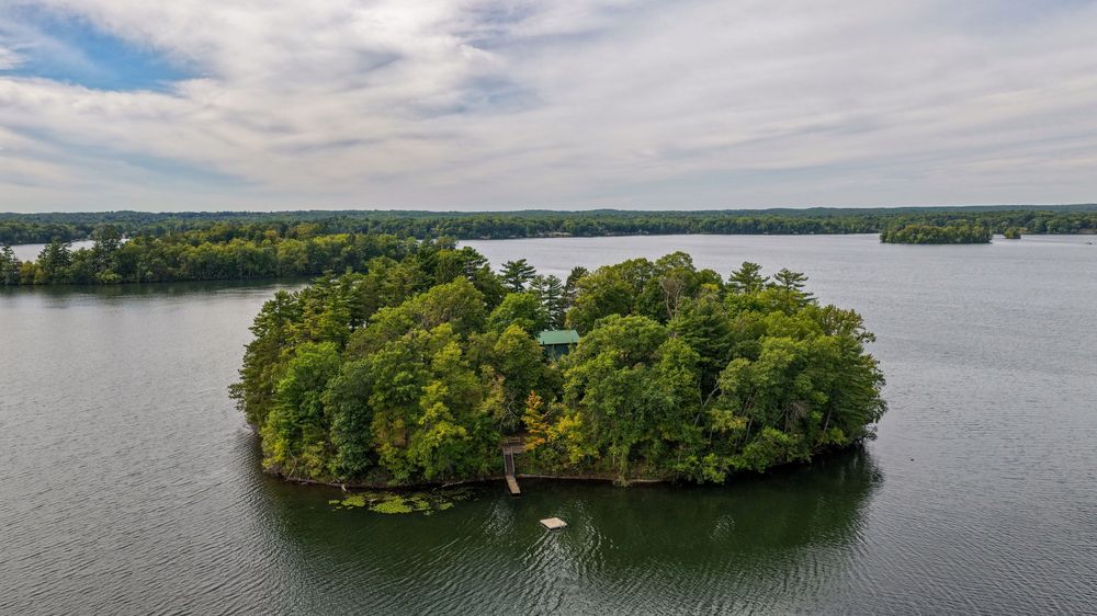 Aerial view of a lush, tree-covered private island surrounded by calm lake waters under a cloudy sky.