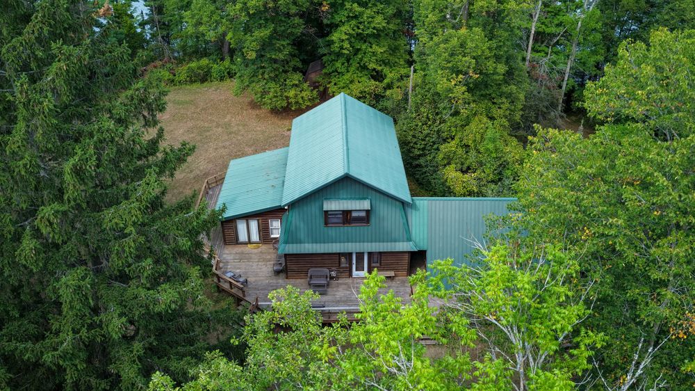 Aerial view of a charming cabin with green metal roof nestled among lush trees on a wooded hillside.