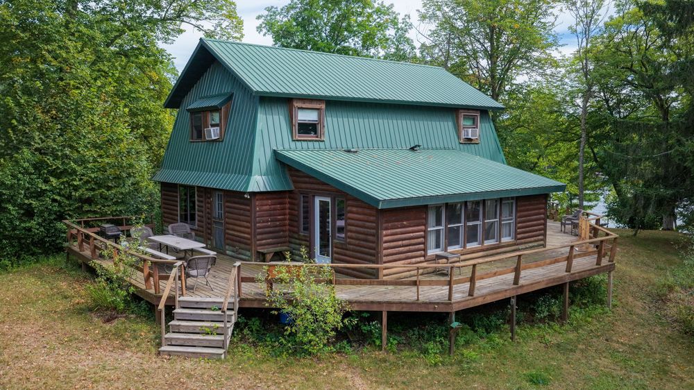 Rustic log cabin with green metal roof and large wooden deck surrounded by lush trees.