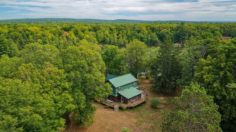 Aerial view of a secluded log cabin with green metal roof surrounded by lush forest trees in a rural wooded setting.