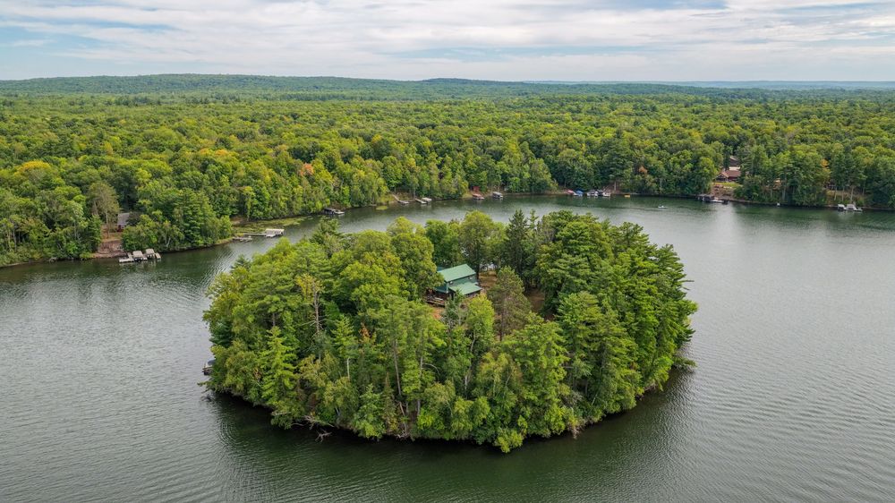 Aerial view of a lush tree-covered island with a cottage surrounded by calm lake waters and dense forest.