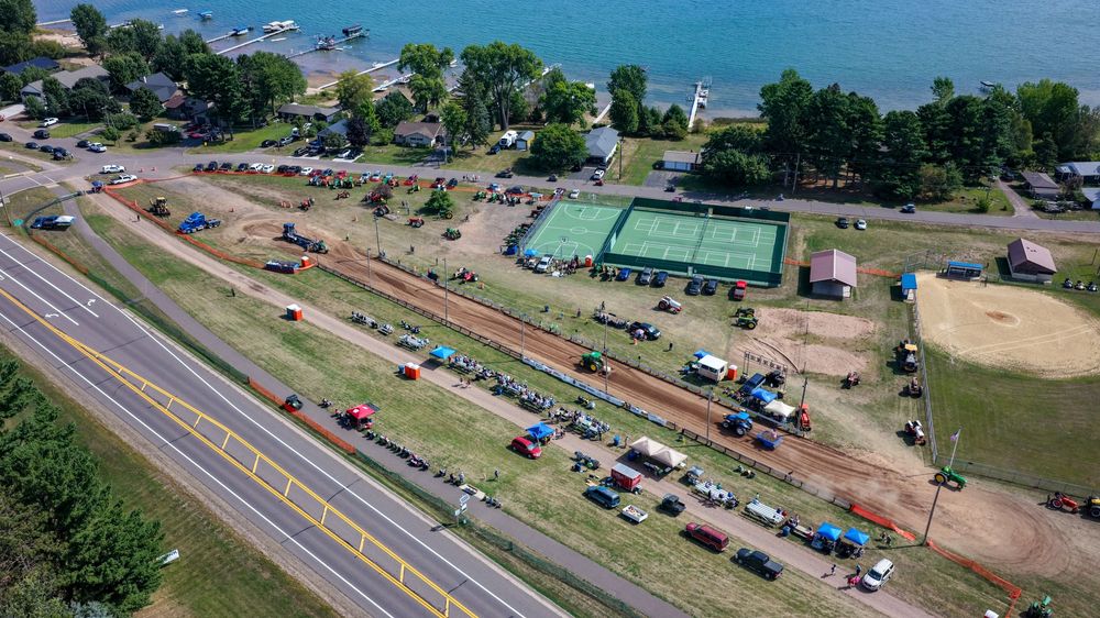 Aerial view of a rural roadside event with tractors, vehicles, and a green tennis court near a lake.