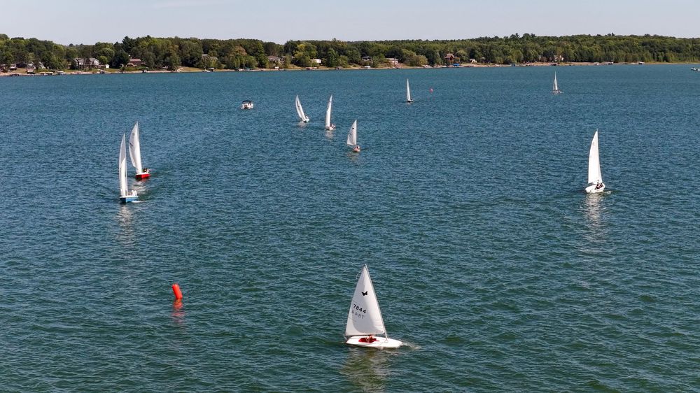 Aerial view of multiple sailboats racing on a calm blue lake surrounded by lush green trees on a sunny day.