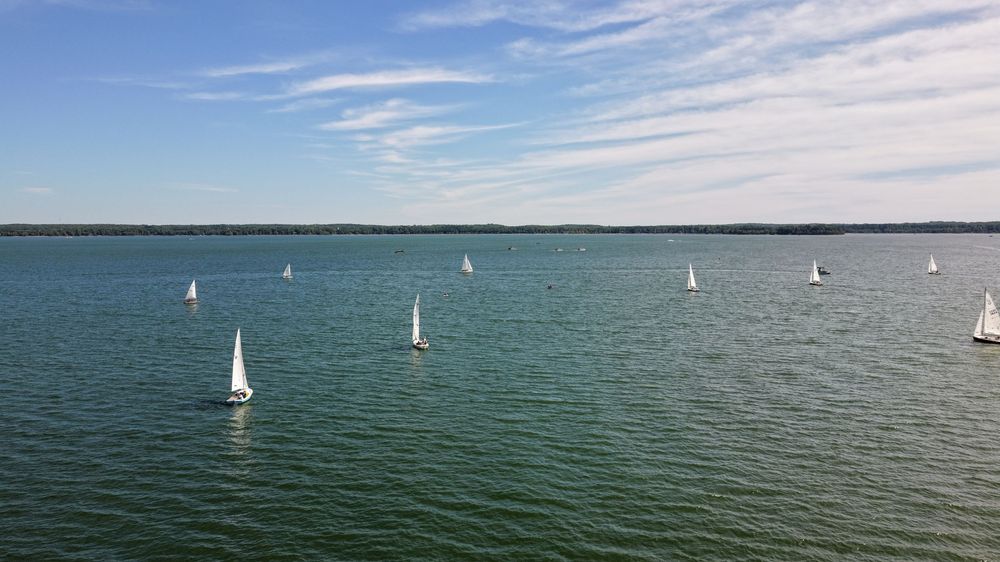 Aerial view of multiple white sailboats gliding across calm blue waters under a partly cloudy sky.